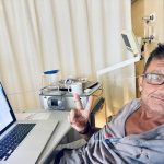 Meuti sitzt optimistisch lächelnd mit einem Peace-Händezeichen im Krankenbett und schreibt den Blogbeitrag